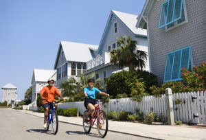 Two people riding bikes down the road past homes with windows